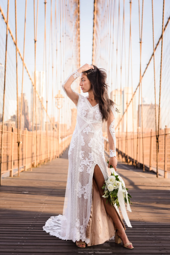 Bridal portrait on the Brooklyn Bridge at golden hour, featuring a fashion-forward lace gown with a high slit and hand-tied bouquet blending New York energy with refined elegance.