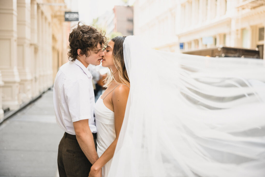 Candid kiss between a bride and groom on a sunlit SoHo street, with the bride’s flowing veil captured mid-air a modern editorial moment full of intimacy and motion.