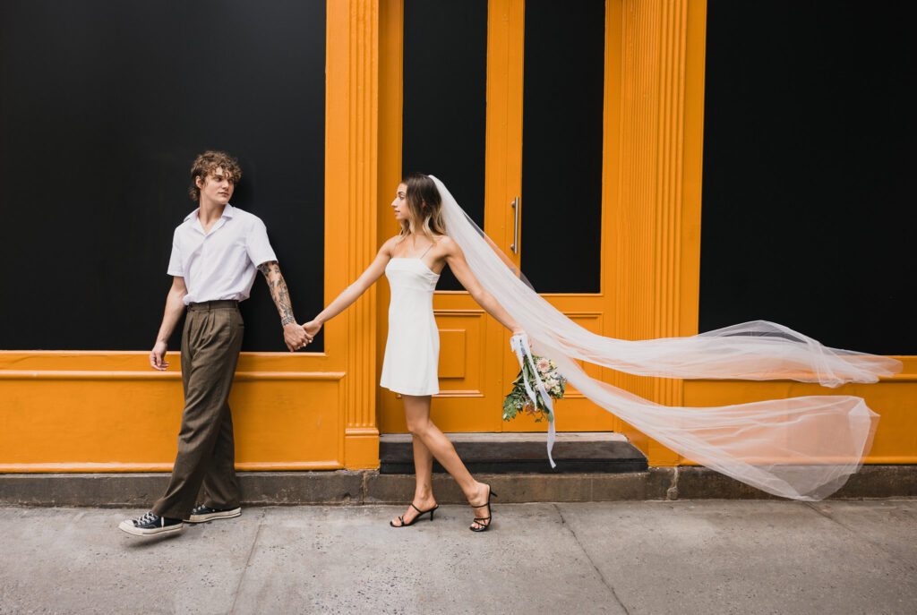 Modern couple holding hands in front of a bold orange storefront in SoHo, with the bride’s veil and bouquet flowing behind her, captured as a chic editorial moment with editorial wedding photography full of movement and style.
