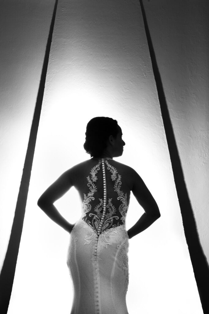 Backlit bridal portrait highlighting the intricate lace detail and buttoned silhouette of a fitted gown, creating a dramatic and artistic editorial composition.