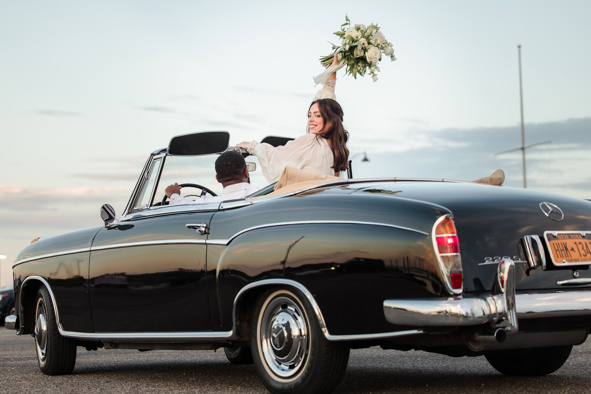 Just married couple drives off in a vintage black Mercedes convertible at sunset, as the bride joyfully raises her bouquet with a smile and looks back over her shoulder.