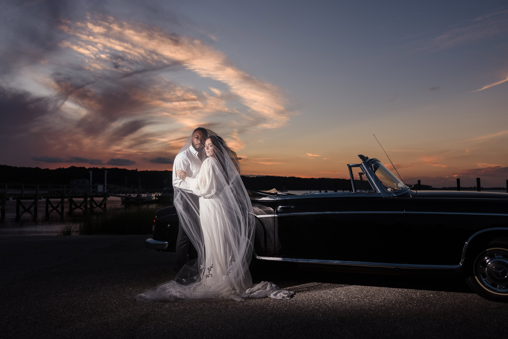 Bride and groom wrapped in a cathedral veil beside a vintage convertible at sunset, with a dramatic sky over the marina — a cinematic editorial portrait blending romance and timeless style.