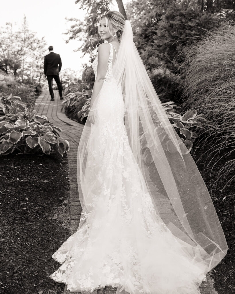 Bride smiling over her shoulder as she walks a garden path, with groom walking ahead in the background.