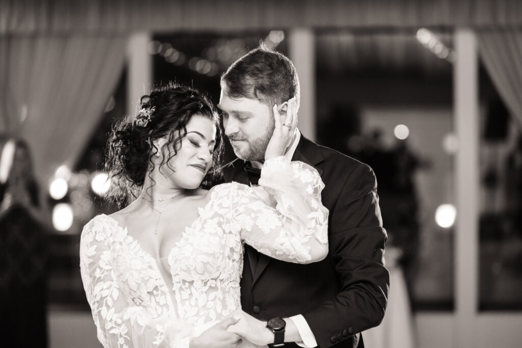 Bride and groom share an intimate first dance in a softly lit reception space, her embroidered gown and his classic tux creating a moment of pure emotion and elegant storytelling, captured with editorial wedding photography.