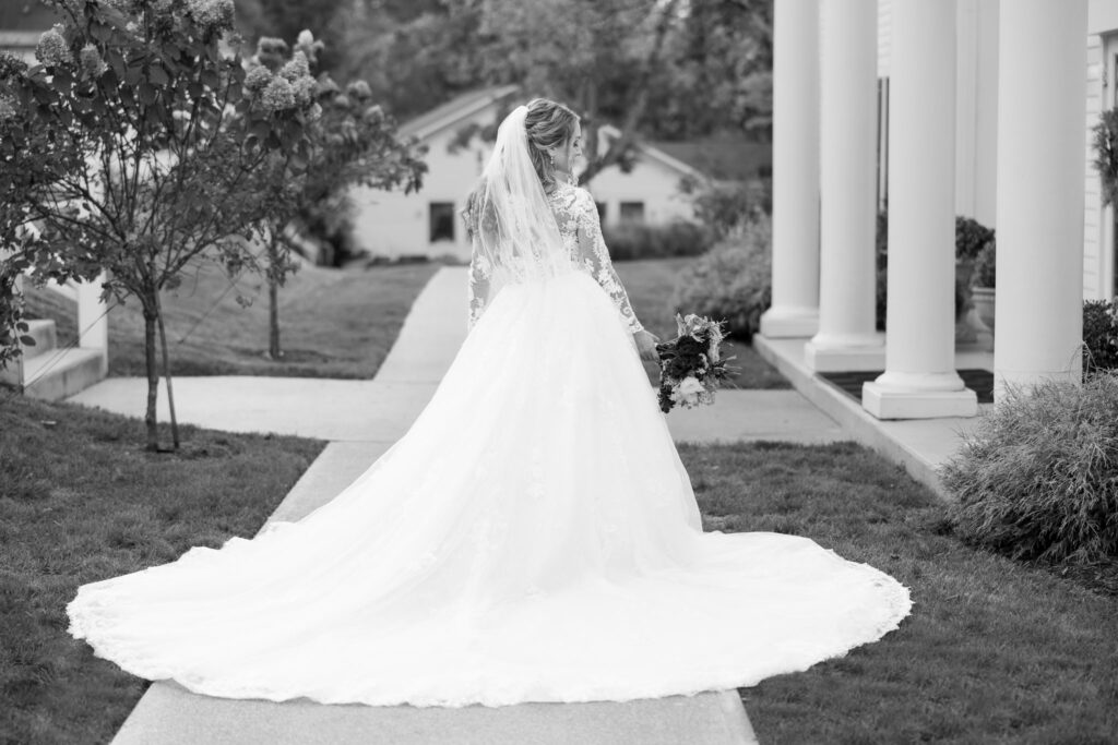 Black and white bridal portrait featuring a bride walking along a garden path with her long lace train flowing behind her, showcasing a classic and timeless wedding photography style.