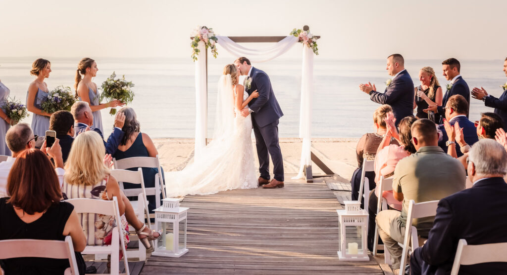 Bride and groom sharing their first kiss under a floral beach arbor during a waterfront wedding ceremony, with guests applauding in soft golden light, captured to illustrate how to choose a wedding photographer with a natural and romantic style.