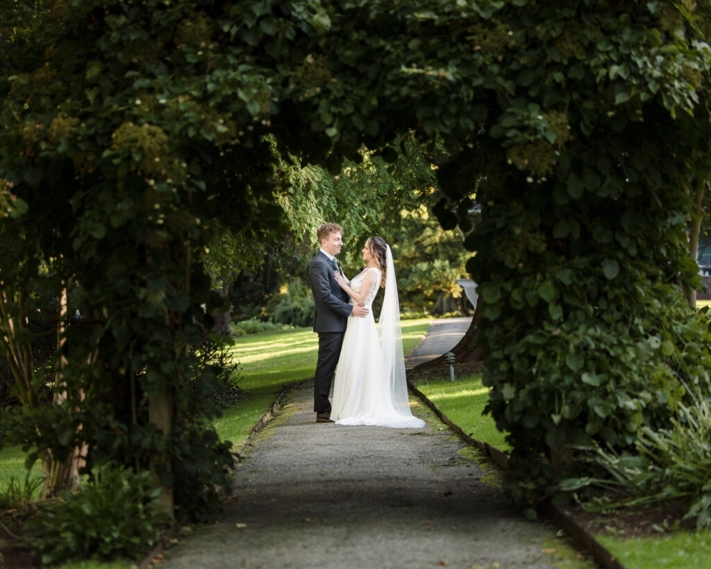 Bride and groom share a quiet, romantic moment beneath a lush garden archway, surrounded by greenery and dappled light, captured as part of premium wedding photography packages.