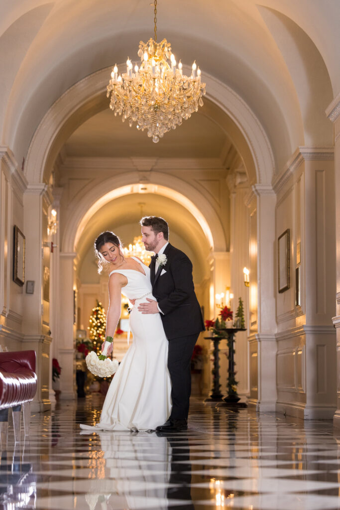 Elegant bride and groom share a romantic moment beneath a grand chandelier in a marble hallway, her minimalist gown and soft pose evoking classic editorial refinement.