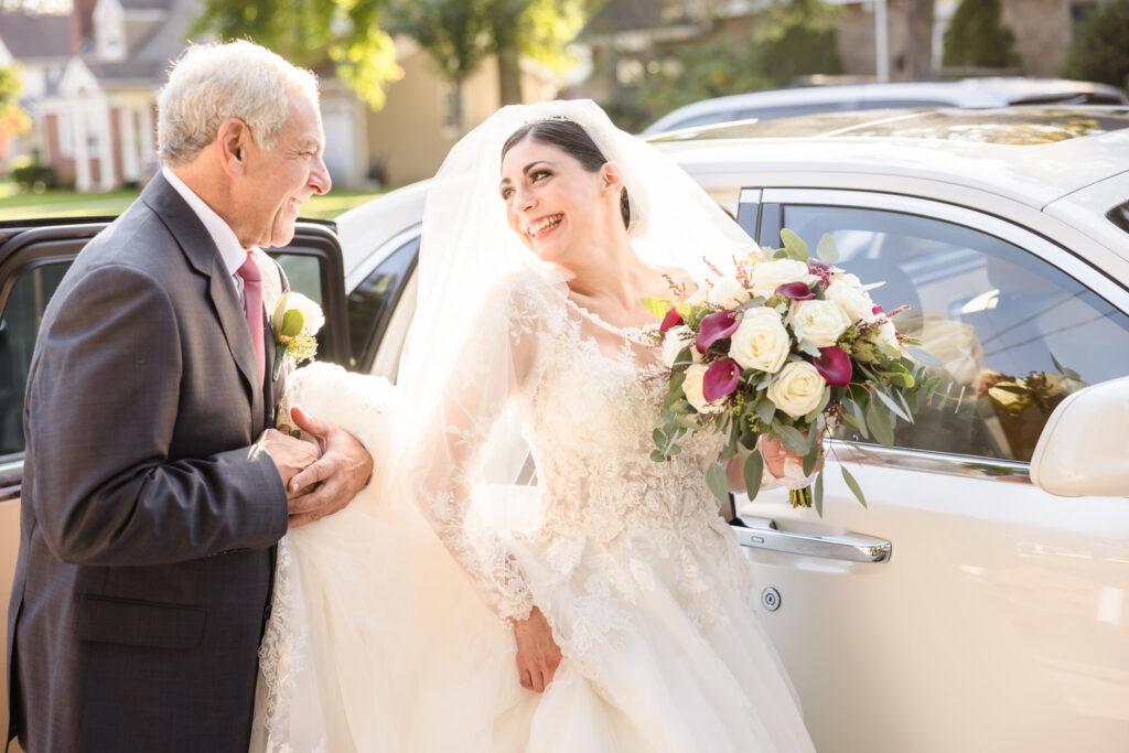 Bride and her father share a joyful, emotional moment as they arrive together for the wedding ceremony, captured candidly outside a white car in golden sunlight.