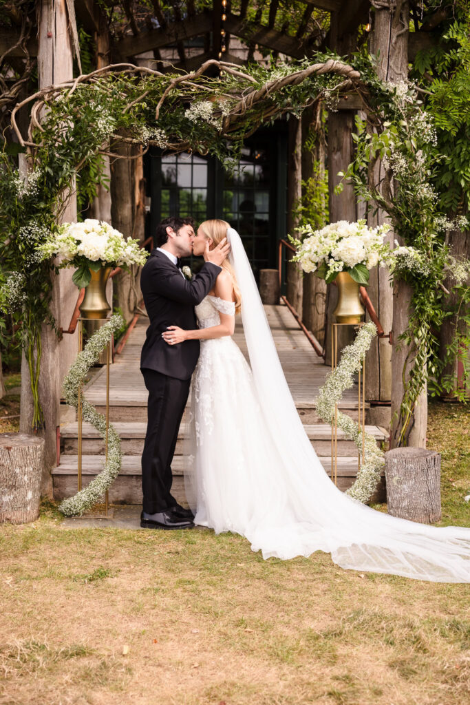 Bride and groom kiss beneath a rustic floral arch at an outdoor ceremony, surrounded by greenery and elegant white florals — a romantic moment of natural beauty and refinement.