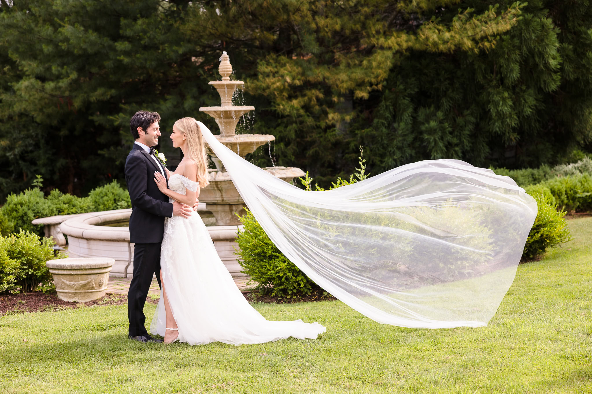 Romantic fine art wedding portrait of a bride and groom embracing in front of a stone fountain, with the bride’s long veil flowing in the wind against a lush garden backdrop.