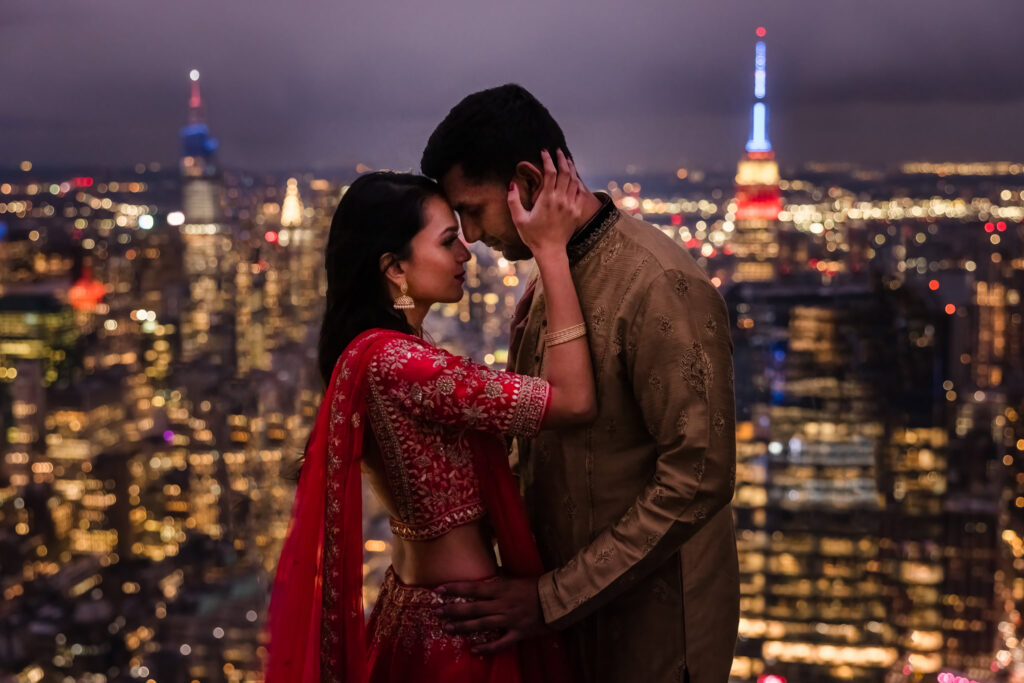 Engaged couple in traditional Indian attire shares an intimate moment atop a New York City skyscraper, with the Empire State Building and glowing skyline behind them.