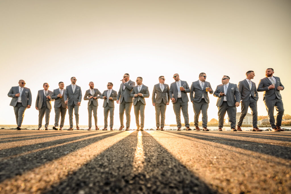 Groom and his groomsmen walk confidently along a sunlit road in matching gray suits, enjoying golden hour light, cigars, and camaraderie before the ceremony.