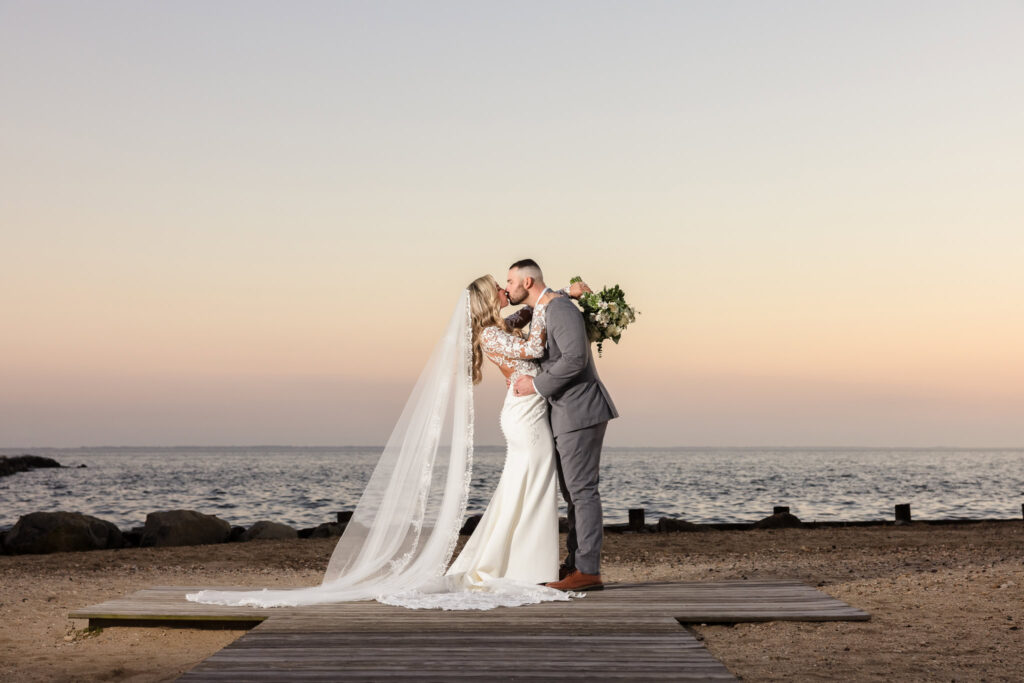 Bride and groom share a kiss on a beach boardwalk at sunset, with a flowing lace veil and soft pastel sky creating a serene and romantic oceanside moment.