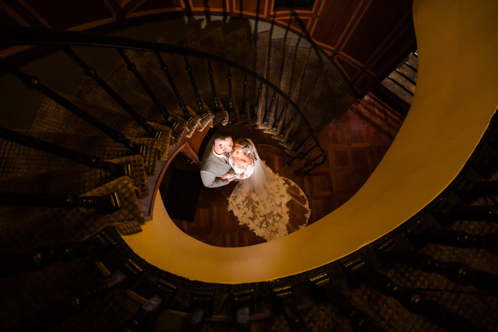 Dramatic cinematic wedding portrait captured from above, showing a bride and groom embracing at the center of a spiral staircase with warm, moody lighting highlighting the bride’s lace train.