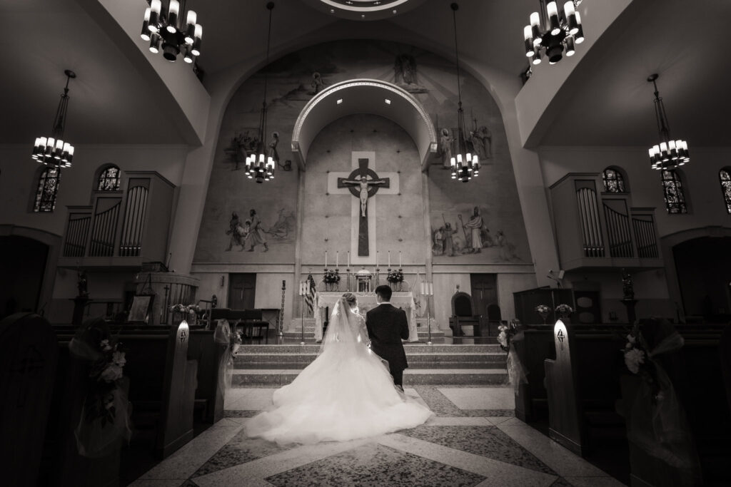 Bride and groom kneeling together at the altar inside a grand cathedral, captured in a dramatic black-and-white style that highlights classic and traditional wedding photography.