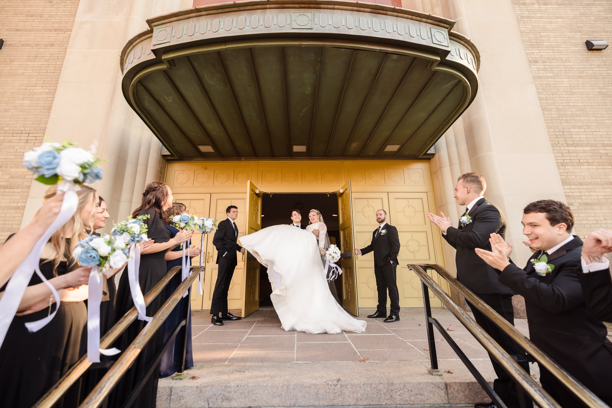 Groom carries his bride through the golden doors of a grand church entrance as their wedding party cheers and waves bouquets — a joyful moment of celebration and tradition.