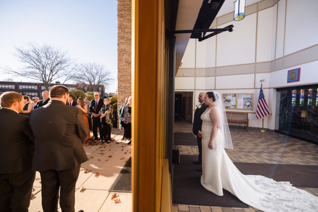 Bride and groom stand inside a church entryway moments before exiting to greet waiting guests, captured through a doorway that frames both their anticipation and the crowd’s excitement.