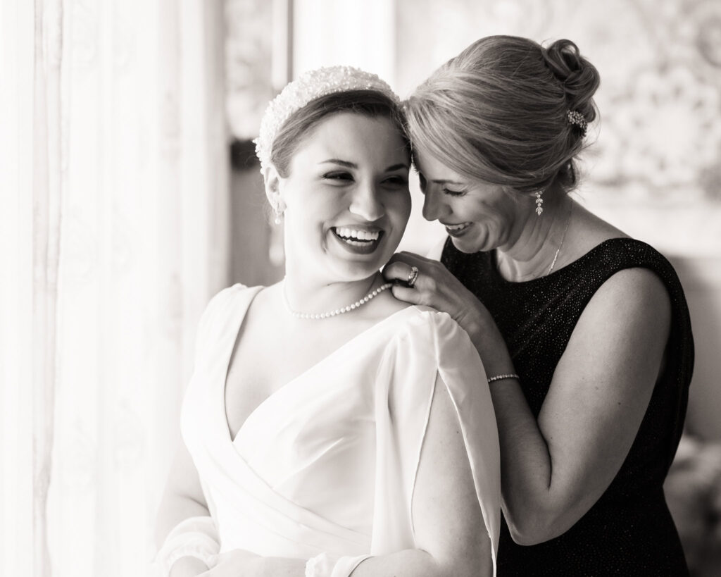 Bride and her mother share a joyful, tearful moment while getting ready, captured in soft window light — a timeless black and white portrait of love and legacy.