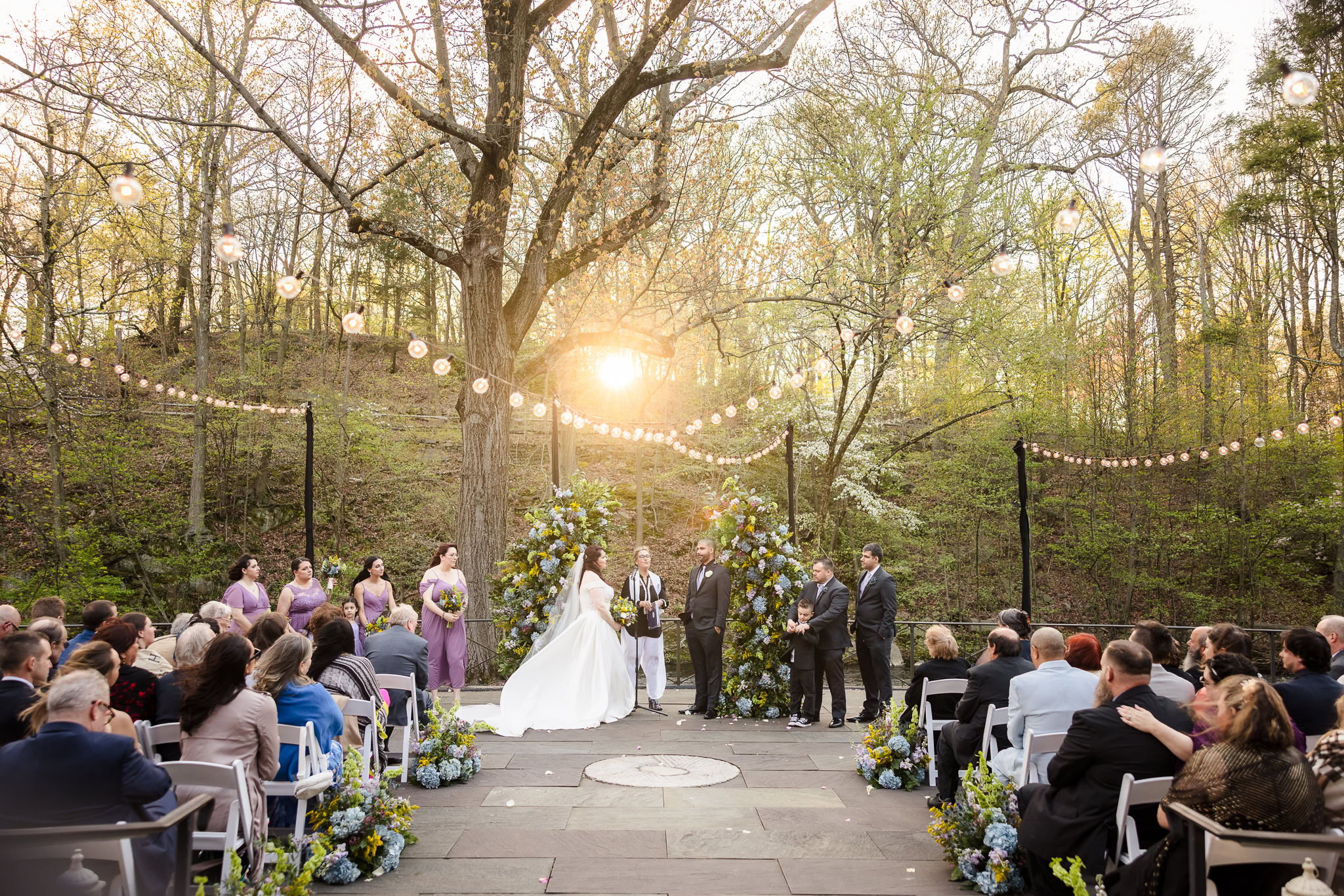 Romantic outdoor ceremony framed by towering trees and golden sunset light, with the couple exchanging vows under string lights and lush floral arches, capturing a cinematic woodland atmosphere.