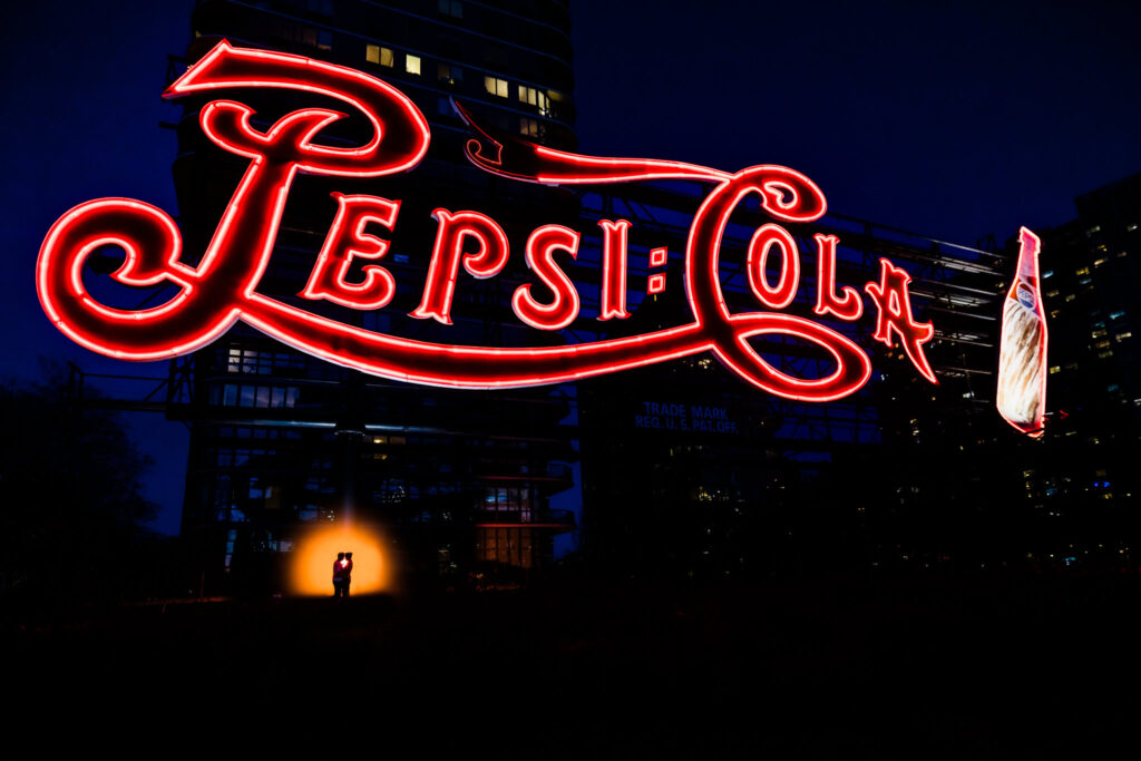 Silhouetted couple embraces beneath the iconic red neon Pepsi-Cola sign in Long Island City, dramatically backlit for a bold, cinematic nighttime portrait, captured with cinematic wedding photography.