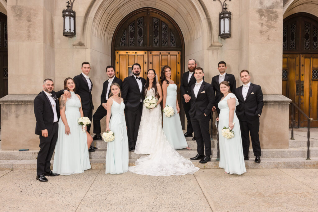 Bride and groom posing with their wedding party in elegant black tuxedos and soft blue dresses outside a grand stone church, highlighting classic and timeless wedding photography style.