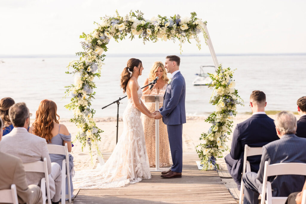 Bride and groom exchange vows under a flower-filled arch during a sunny beachfront wedding ceremony, captured while considering wedding photography cost.