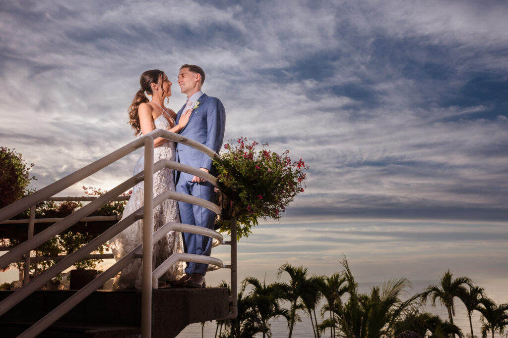 Bride and groom sharing a romantic moment on a staircase at sunset with dramatic skies and palm trees, highlighting artistic and cinematic wedding photography style.