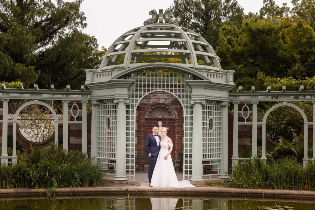 Bride and groom standing beneath an ornate garden pavilion with classical architecture, captured in a timeless and elegant wedding portrait style.