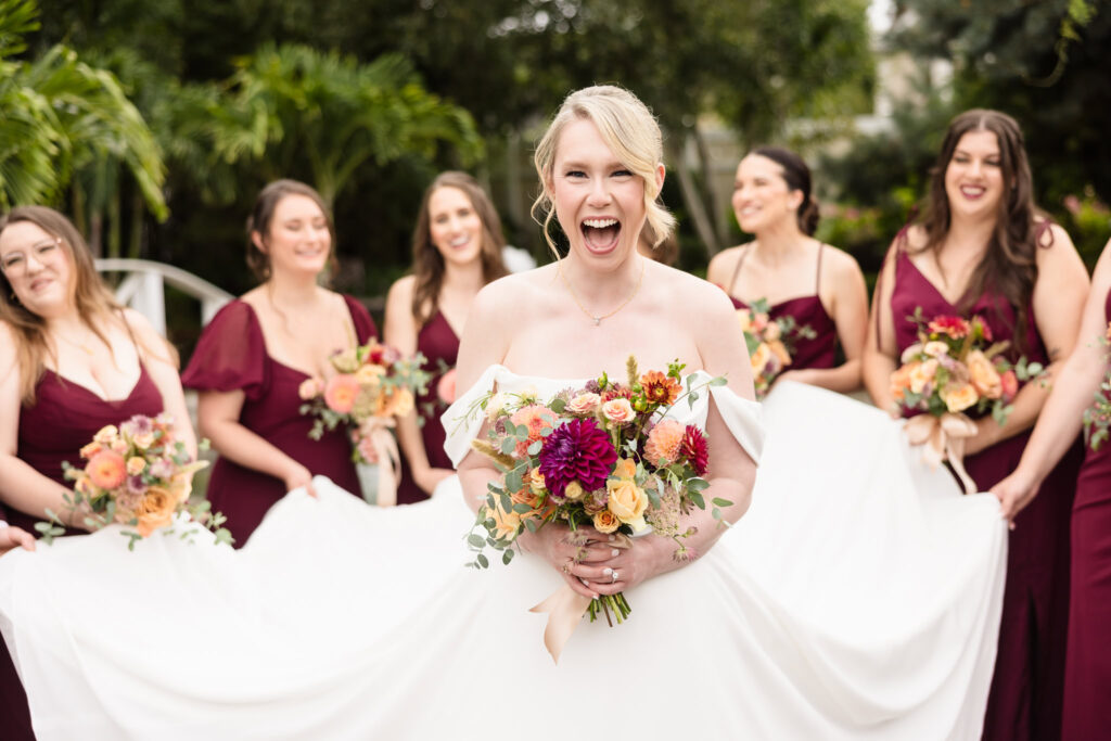 Bride laughing joyfully while holding a colorful bouquet, surrounded by bridesmaids in burgundy dresses during an outdoor wedding portrait.
