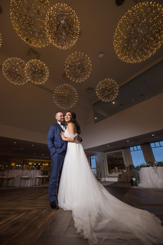 Bride and groom embrace beneath golden chandeliers in a modern ballroom, her long train trailing across the wood floor — a refined portrait of elegance and joy.