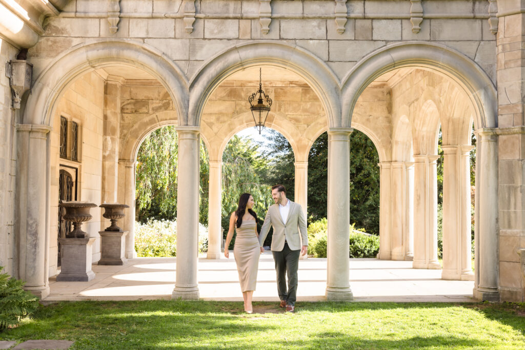 Engaged couple walking hand in hand beneath elegant stone archways in a garden courtyard, showcasing timeless and sophisticated engagement photography.