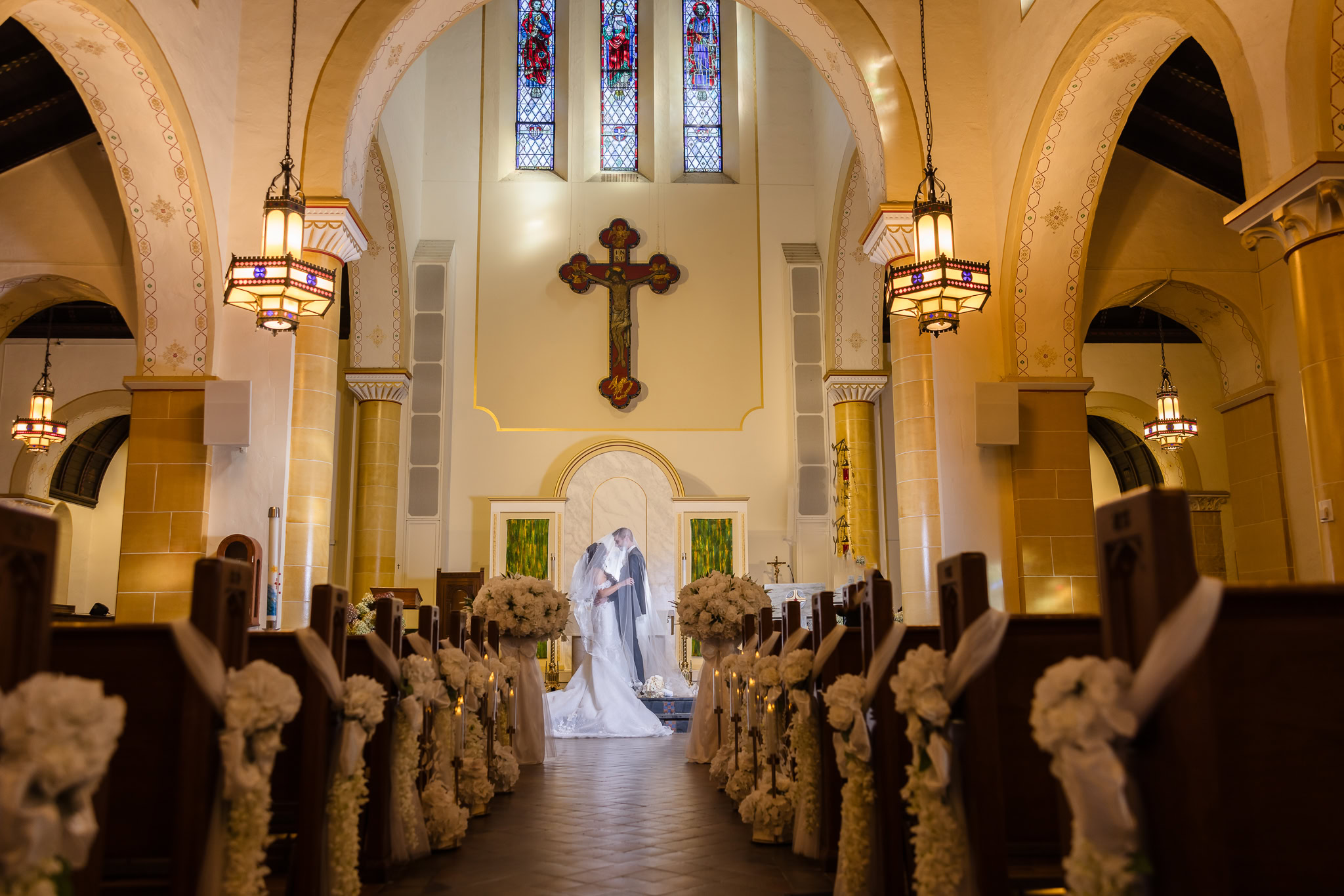 Bride and groom sharing a kiss at the altar of an ornate church, surrounded by floral aisle décor, photographed in a traditional wedding style.