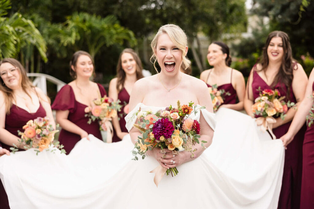 Bride laughing joyfully while holding a vibrant fall bouquet, surrounded by bridesmaids in deep burgundy dresses, captured to illustrate how to choose a wedding photographer with natural, candid wedding photography style.