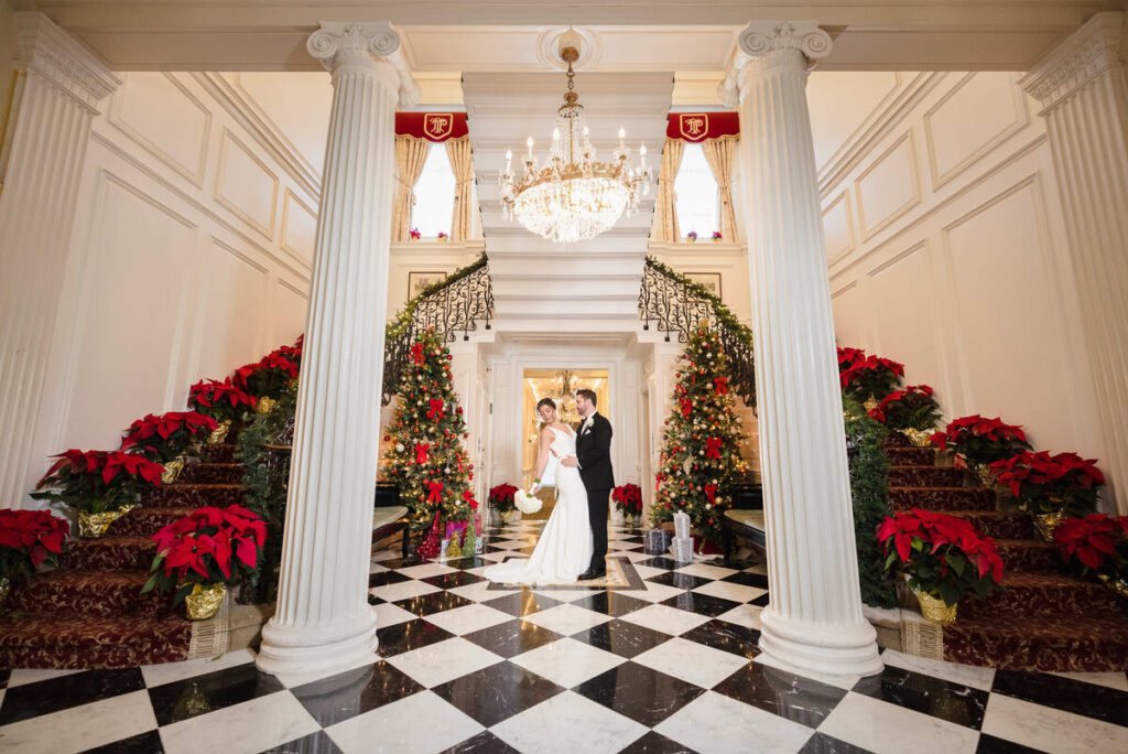 Bride and groom posing in an elegant mansion foyer decorated for Christmas, with grand staircases, poinsettias, and glowing holiday trees creating a luxurious wedding portrait backdrop an image style often searched on the best platforms to compare wedding photographers.