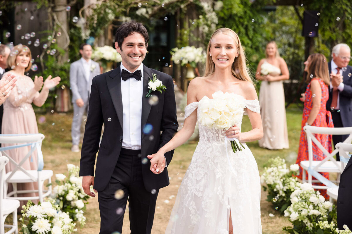 Bride and groom walk joyfully down the aisle after their ceremony, surrounded by smiling guests and soft bubbles in a garden full of white florals and greenery.
