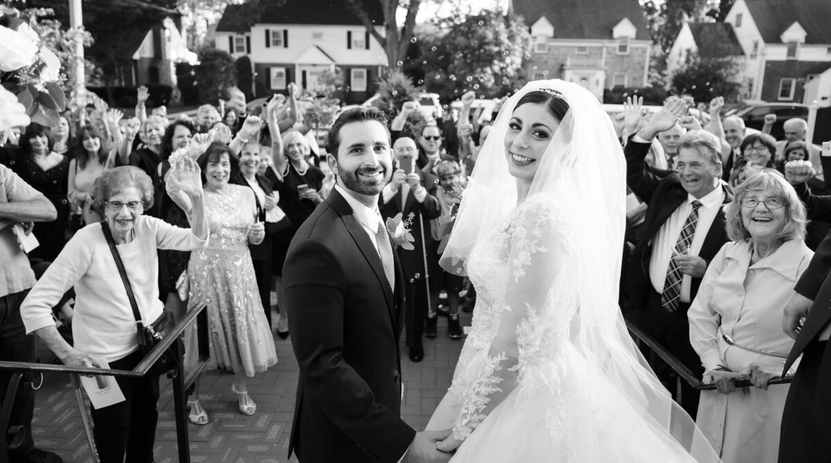 Bride and groom smiling as they walk out of their ceremony while guests cheer and celebrate around them, captured in a classic black-and-white documentary wedding photography style.