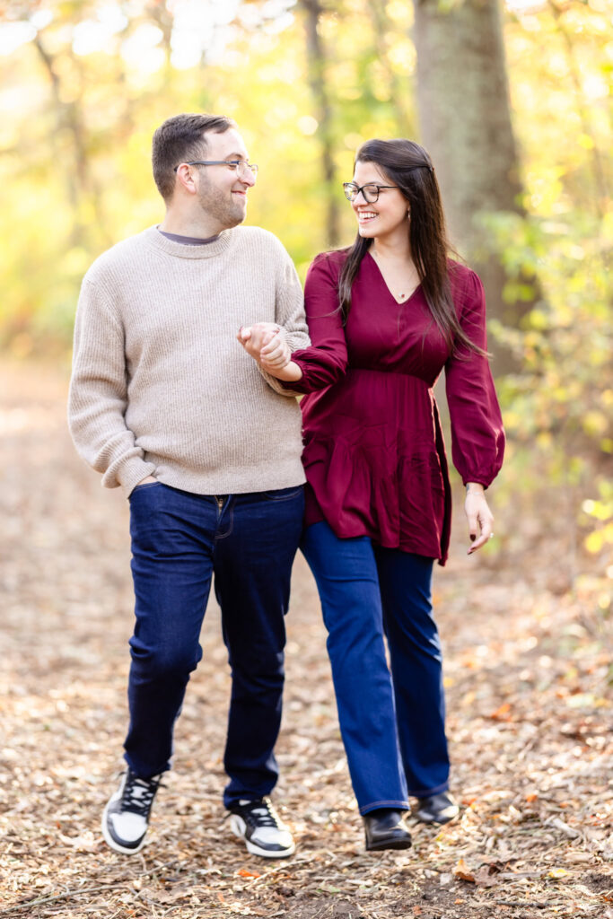 Engaged couple walking hand in hand through a sunlit wooded trail at Bethpage State Park during their fall engagement session.
