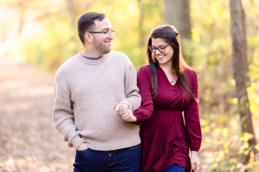 Smiling couple walking arm in arm through a golden forest path during their fall engagement session at Bethpage State Park.