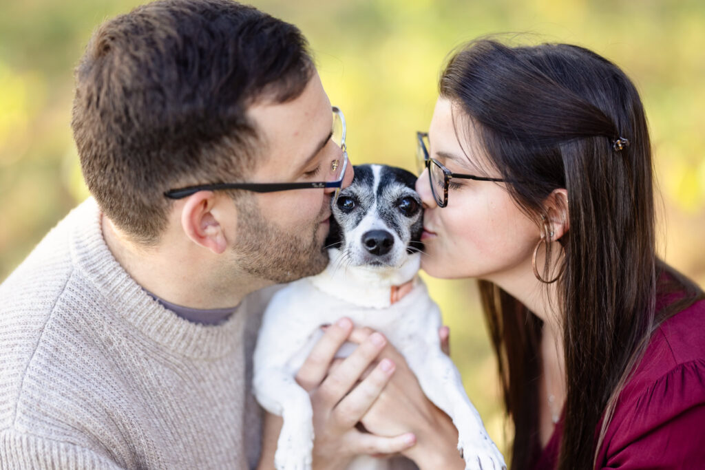 Engaged couple giving their dog a kiss on each cheek during their fall engagement session.