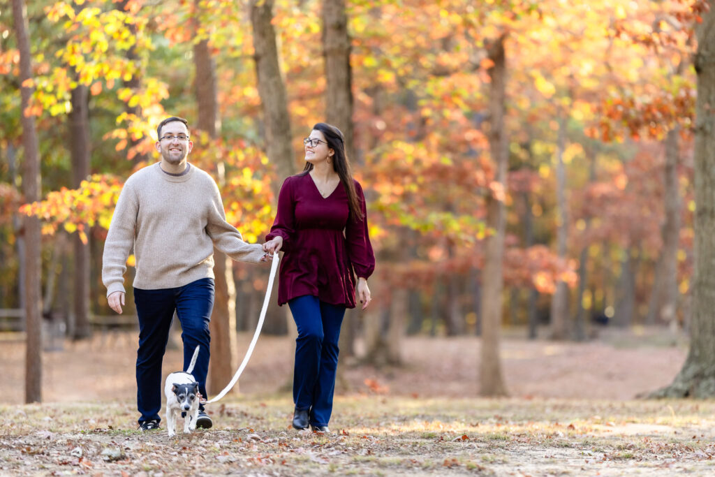 Couple walking their dog through a forest trail lined with vibrant autumn leaves during their Bethpage State Park engagement session.