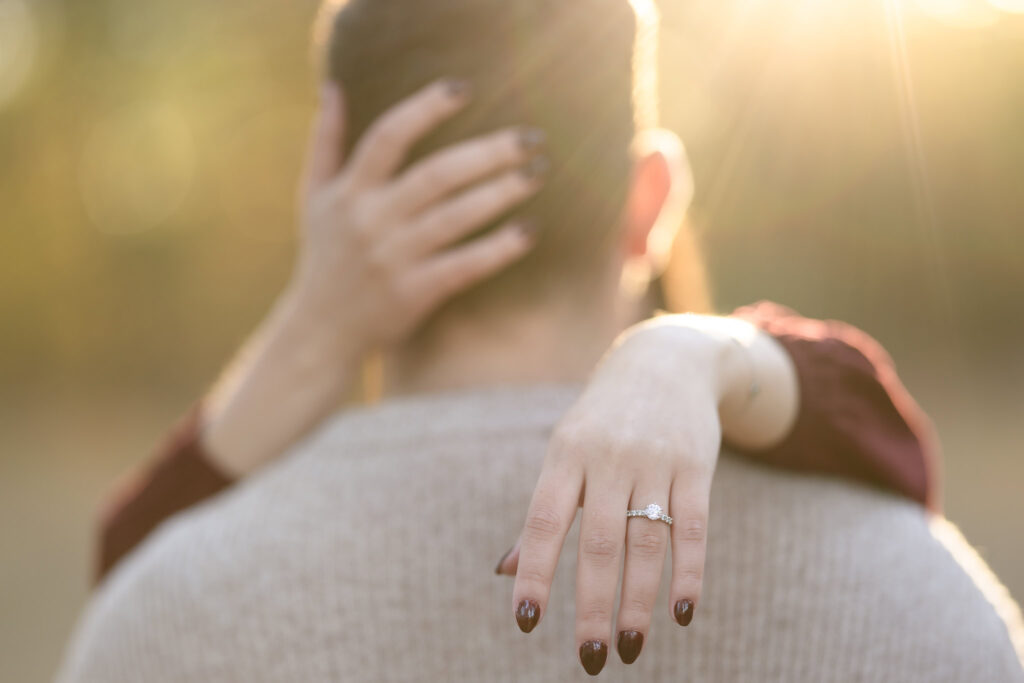 Close-up of engagement ring on bride-to-be’s hand resting on her fiancé’s shoulder, glowing in the golden sunset at Bethpage State Park.