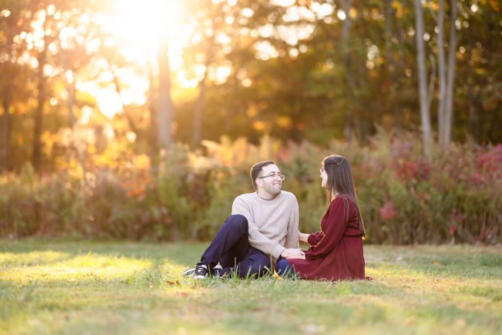 Couple sitting in a sunlit meadow surrounded by autumn foliage, sharing a quiet moment during their Bethpage State Park engagement session.