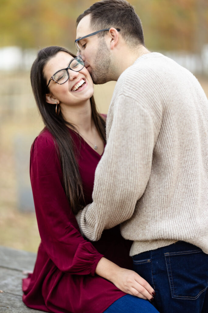 Groom-to-be kissing his fiancée on the cheek as she laughs joyfully during their fall engagement session at Bethpage State Park.