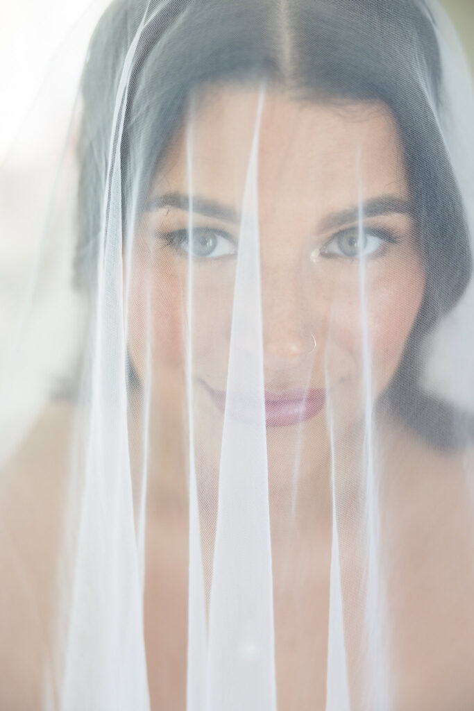 Bride smiling softly behind her sheer wedding veil in a romantic bridal portrait before her ceremony.