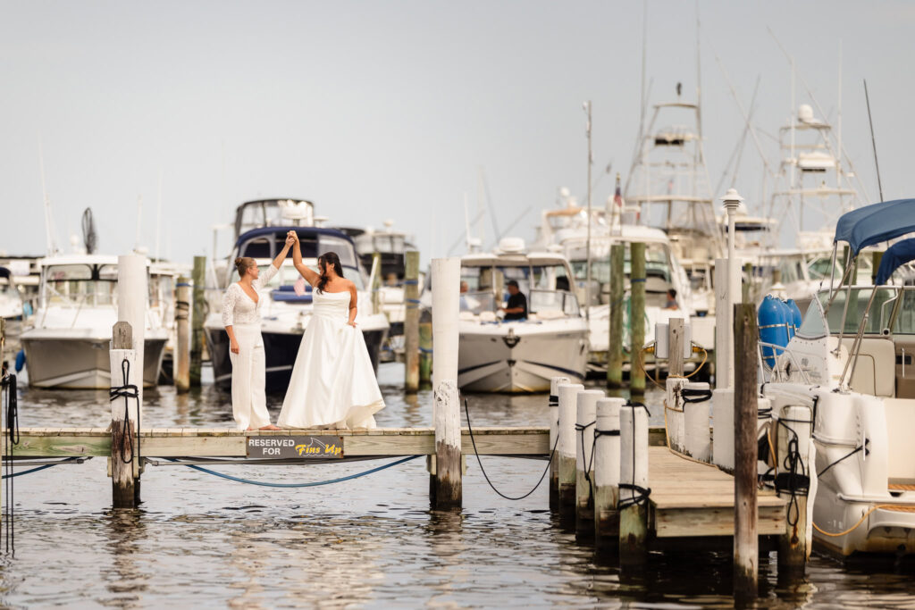 Brides dancing together on a dock surrounded by boats at the Snapper Inn marina during their waterfront wedding portraits.