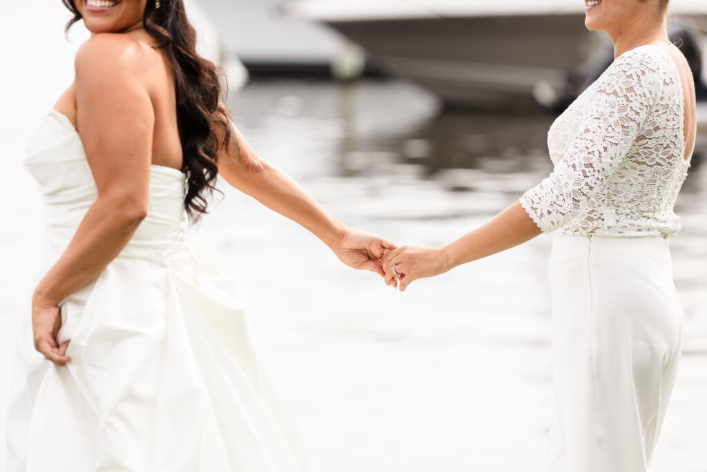 Close-up of two brides holding hands during waterfront wedding portraits at The Snapper Inn, featuring elegant lace and satin attire.