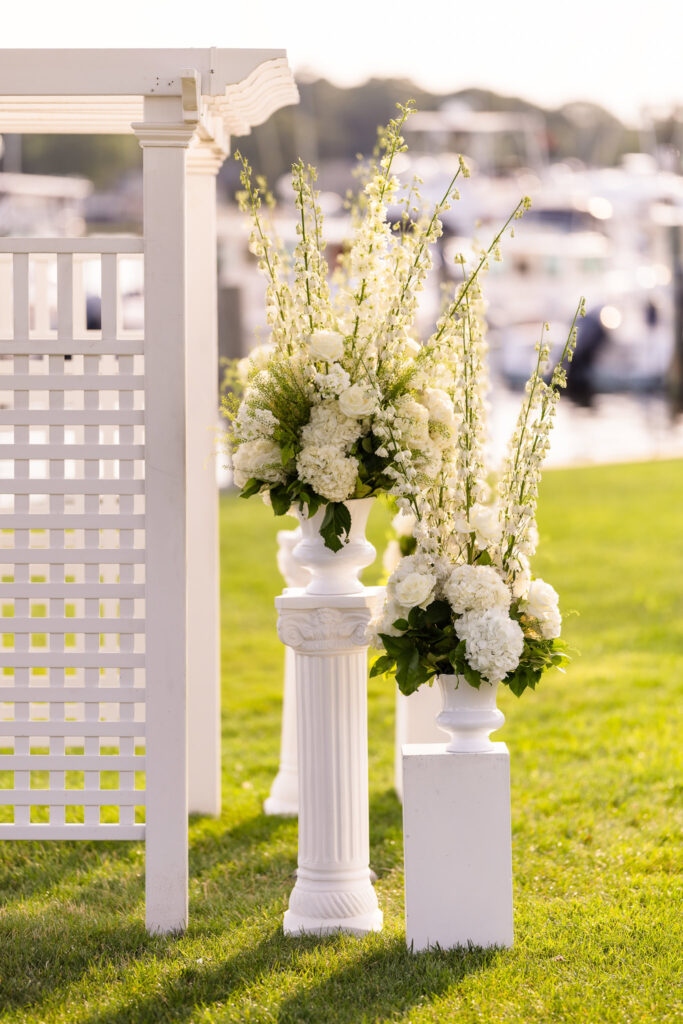 White floral arrangements with hydrangeas and roses displayed on classic columns at a sunlit Snapper Inn waterfront ceremony.