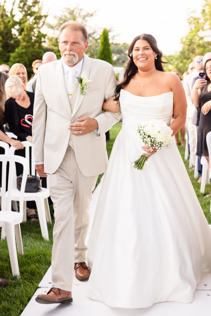 Bride walking down the aisle with her father during an outdoor waterfront ceremony at The Snapper Inn in Oakdale.