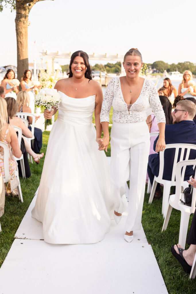 Newly married brides walking up the aisle hand in hand, smiling joyfully after their waterfront ceremony at The Snapper Inn.